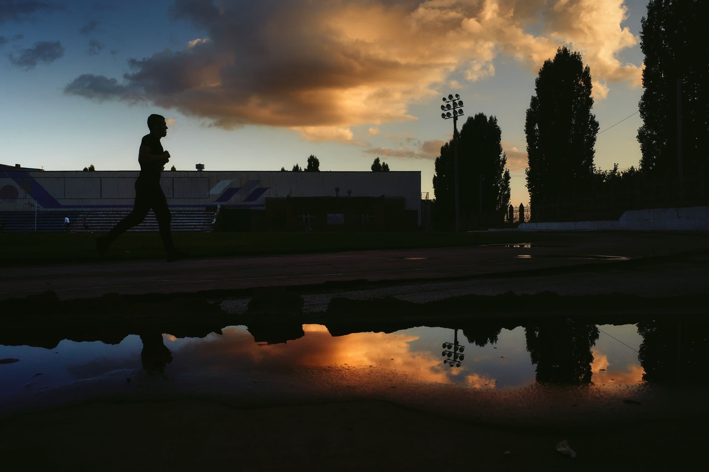 Silhueta de um corredor no estádio ao entardecer, com o céu refletido numa poça
